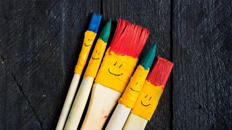 Five paintbrushes with different colored bristles and smiley faces on their handles are arranged on a wooden surface.