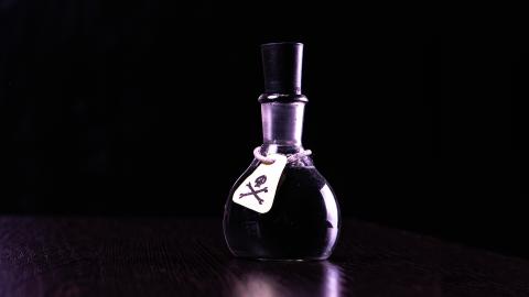 A small dark vial with a cork top and a skull and crossbones warning label on a wooden surface against a black background.