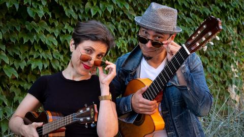 Two people with sunglasses, one holding a ukulele and the other a guitar, posing in front of a leafy background, looking cool.