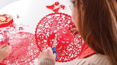 A person crafting intricate red paper cutouts with scissors on a white table, with red tassels and envelopes nearby.