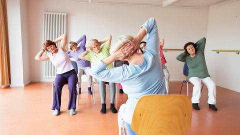 A group of elderly people are seated on chairs, stretching with their hands behind their heads in a bright room.