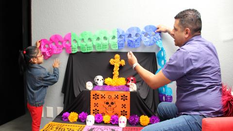 Father and daughter decorate an ofrenda with colorful papel picado and skulls for Día de los Muertos.