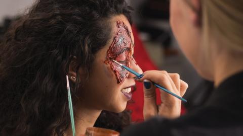 Person applying special effects makeup that mimics wounds to another person's face in a professional setting.