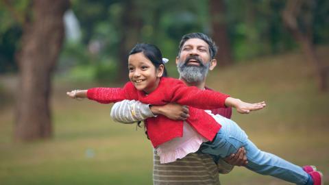 An older man holds a smiling young girl in an outstretched "flying" pose in a park. Both look joyful.