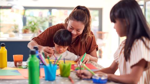 A mother helps her young child with a painting project, while an older child paints at the table in a bright, cozy room.