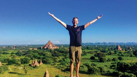 A person with arms outstretched stands on a ledge overlooking a lush landscape with pagodas on a sunny day.