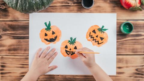 Child painting jack-o'-lantern faces on paper with orange paint, surrounded by art supplies and a pumpkin on a wooden table.