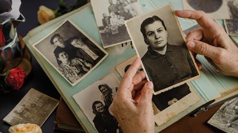 Hands holding an old photo of a woman, surrounded by other vintage photographs and a scrapbook on a table.