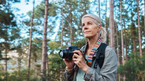 A woman with a camera stands in a forest, gazing thoughtfully at the surroundings.