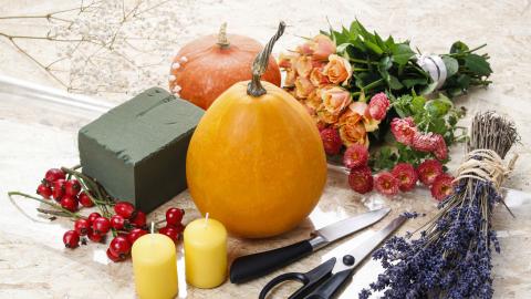 A table with pumpkins, flowers, candles, scissors, and a floral foam block arranged for a fall-themed craft project.