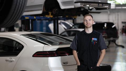 Mechanic in a black uniform stands in an auto repair shop with cars on lifts in the background.