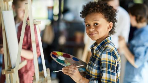 A child smiles while painting on a canvas. Other people are painting in the background.