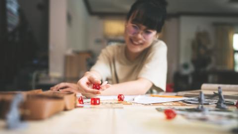 Person playing a tabletop role-playing game, rolling red dice on a cluttered game board, smiling and leaning forward.