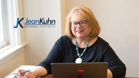 Woman with glasses smiling, seated at a table with a laptop, near a window, wearing a black top and turquoise necklace.