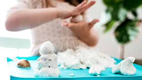 Child shaping white dough on a blue tray, with a spoon and other dough shapes visible. Green plant blurred in background.