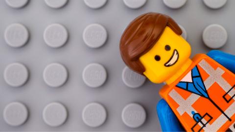 A LEGO minifigure with brown hair, a happy face, and an orange construction outfit lying on a grey LEGO baseplate.