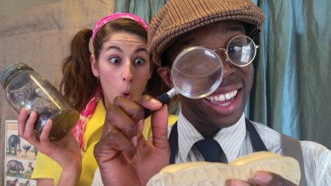 Two people smiling, one holding a magnifying glass to a model, the other holding a jar, in a playful setting.