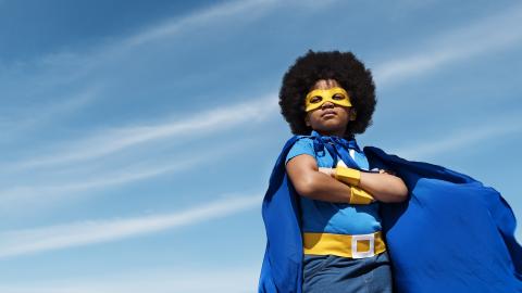 A child wearing a superhero costume with a blue cape and yellow mask stands confidently against a blue sky.