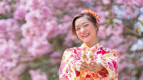 Smiling woman in floral kimono holds flowers against a backdrop of pink blossoms.