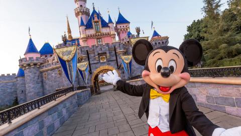 Mickey Mouse poses in front of Sleeping Beauty Castle at Disneyland, dressed in his classic red shorts and yellow bow tie.