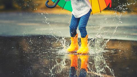 Child wearing yellow rain boots jumps in a puddle, holding a colorful umbrella. Splashes of water around.