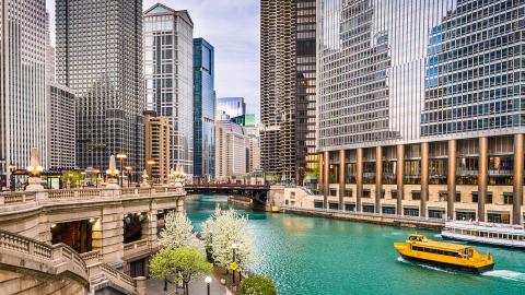 Chicago River with a water taxi, surrounded by tall buildings and blooming trees along the riverwalk under blue skies.