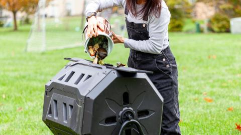 Person emptying a bucket of food scraps into a black compost bin outdoors.