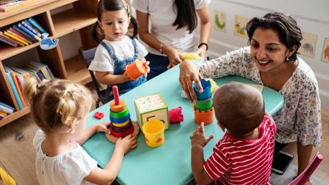 Children and a teacher playing with stacking toys at a small table in a classroom setting.