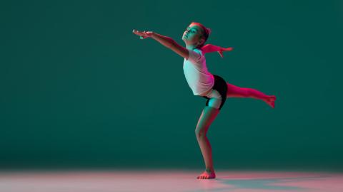 Young girl in athletic wear gracefully balancing on one leg against a teal background.