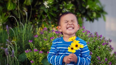 A child in a blue striped shirt smiles with eyes closed, holding yellow flowers, standing in a garden with purple blooms.