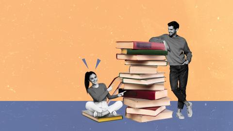 A woman sitting and pointing at a tall stack of books, with a man standing beside the pile, both smiling.