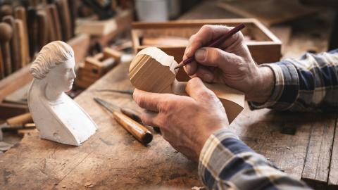 Close-up of hands carving a wooden bust in a workshop, with tools and a reference sculpture nearby.