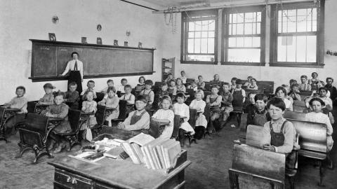 Black-and-white photo of a vintage classroom with children seated at desks and a teacher by the chalkboard.