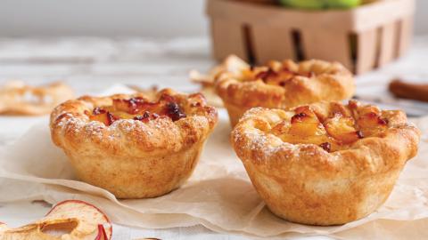 Three small apple pies with golden crusts on parchment paper, with a basket and dried apple slices in the background.