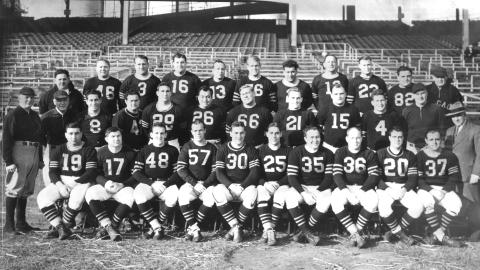 Black-and-white photo of a vintage football team posing in uniforms and striped socks in a stadium.