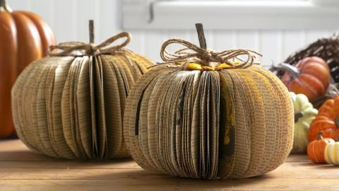 Two pumpkins made from folded book pages, each tied with twine stems, on a wooden surface with other autumn decorations.