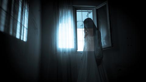 A ghostly figure in a white dress stands by a window with sheer curtains in a dimly lit room.