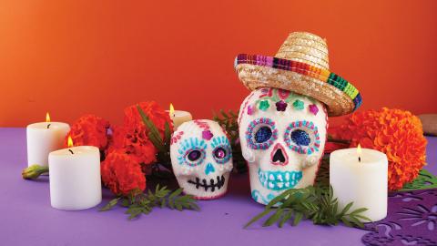 Colorful Day of the Dead setup with decorated sugar skulls, flowers, and a vase of marigolds against a yellow and orange background.