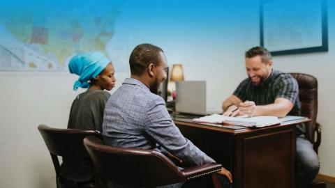 A couple sits across a desk from a smiling man in an office, discussing paperwork.