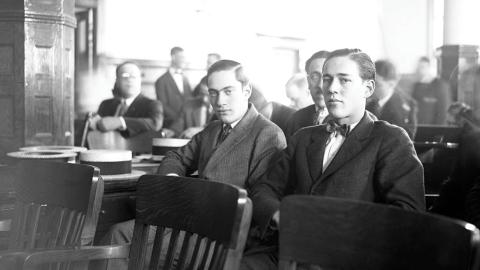 Two young men in suits sit in a courtroom with others visible in the background.