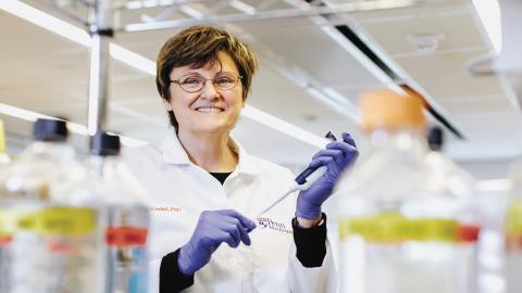 A scientist in a lab coat and gloves holds a pipette, smiling among bottles in a laboratory.
