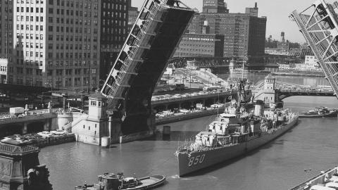 A navy ship marked "850" passes under an open drawbridge in a city river, with tall buildings nearby.