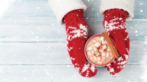 Hands in red mittens hold a mug of hot chocolate with marshmallows and a cinnamon stick, surrounded by snow.