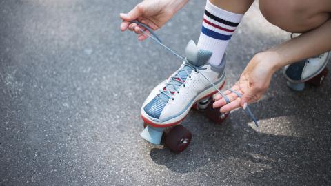 Person tying the shoelaces of a white roller skate on an asphalt surface, wearing striped socks.