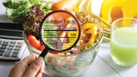 A hand holds a magnifying glass over a salad bowl, highlighting the nutrition facts label showing 120 calories.