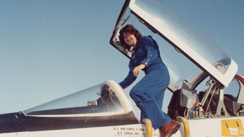 A woman in a blue jumpsuit climbs into the cockpit of a U.S. Air Force jet under a clear sky.