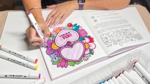 A person colors a floral heart design in an adult coloring book with bright markers on a table.