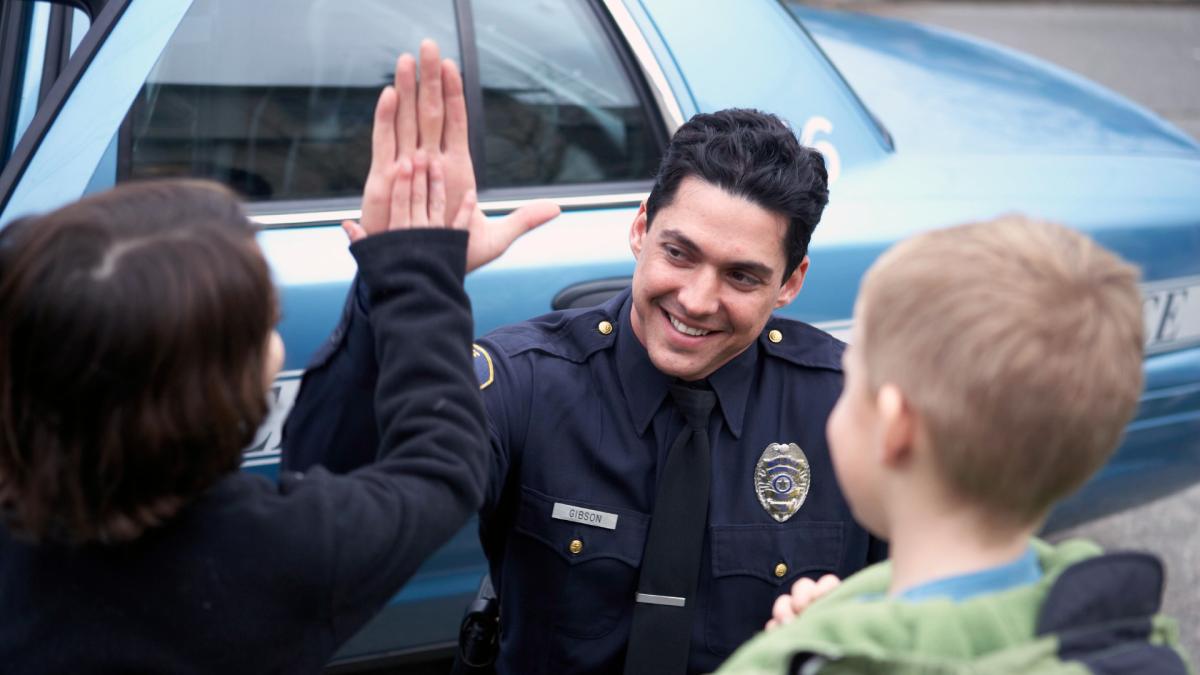 Police officer giving a high-five to a child while another child watches, standing next to a patrol car.