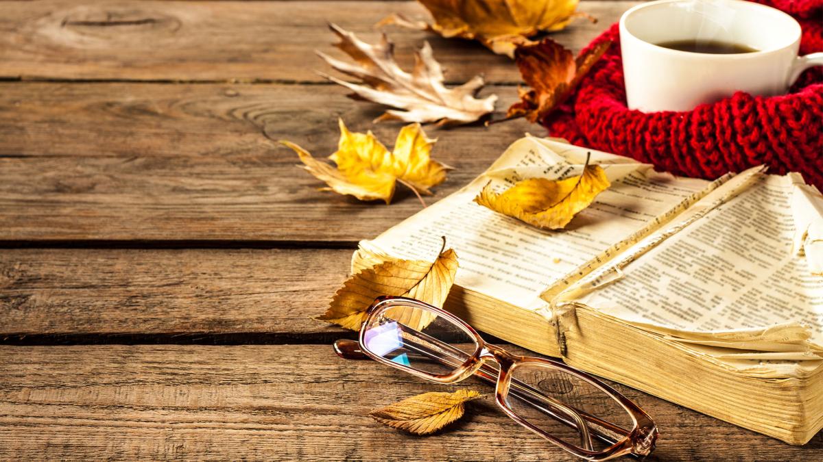 Open book, glasses, autumn leaves, and a coffee cup wrapped in a red scarf on a wooden table.