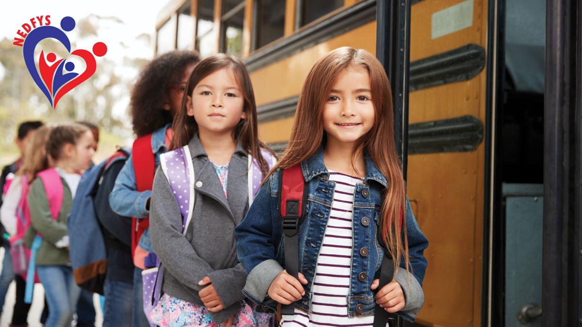 Children in a line, smiling, as they board a yellow school bus. They are wearing backpacks and casual clothing.